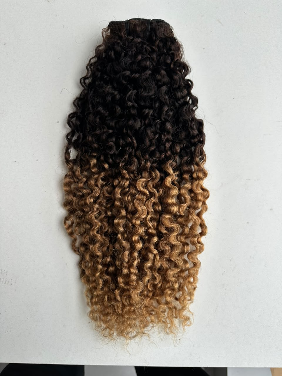 Curly hair weave extension with a dark brown to golden blonde ombré color transition, shown against a plain white background.