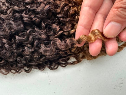 Curly hair extension or wig section with a dark brown to golden blonde ombré color, held by a hand against a light gray background.